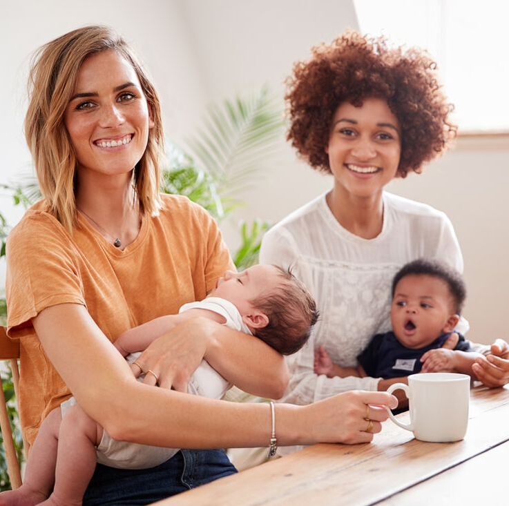 Two mothers holding babies and smiling over coffee. Feeling confident about their postpartum season and transition into motherhood with support in group therapy. Help is available in Ewing, NJ for pregnant and postpartum mothers.